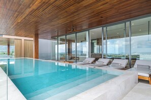 Una piscina interna moderna con acqua cristallina, soffitto in legno e ampie vetrate che offrono vista all'esterno.