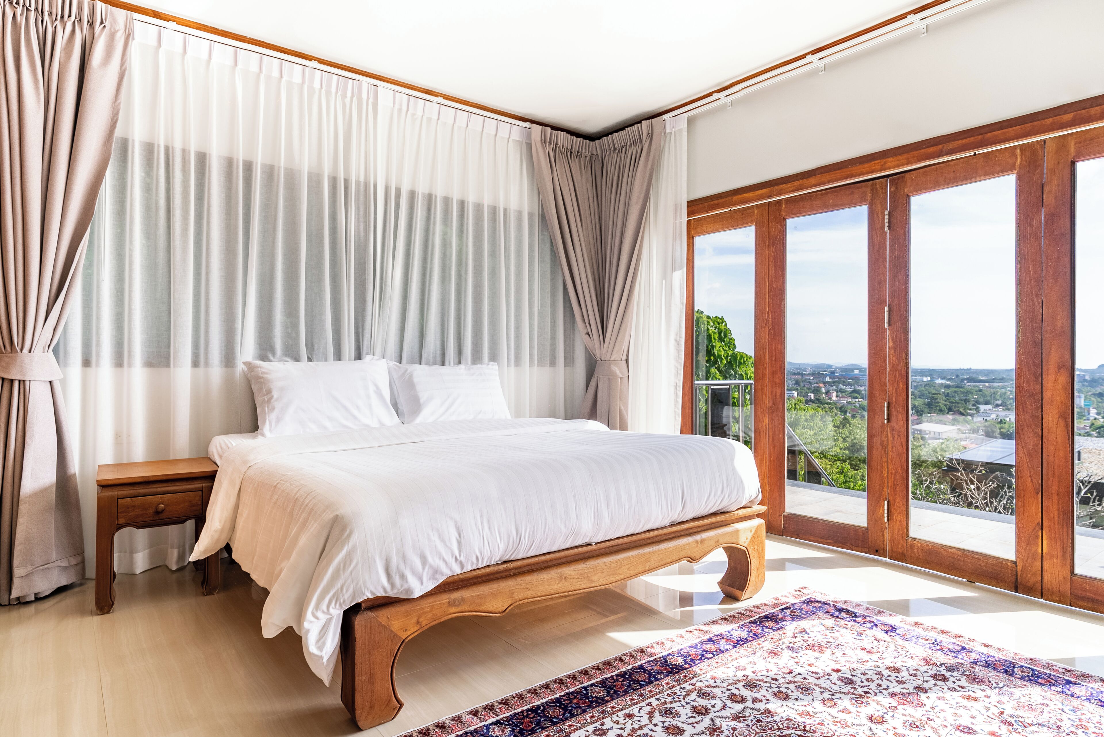 A bedroom with a large bed, a wooden nightstand, a view through a sliding glass door, and a patterned rug.