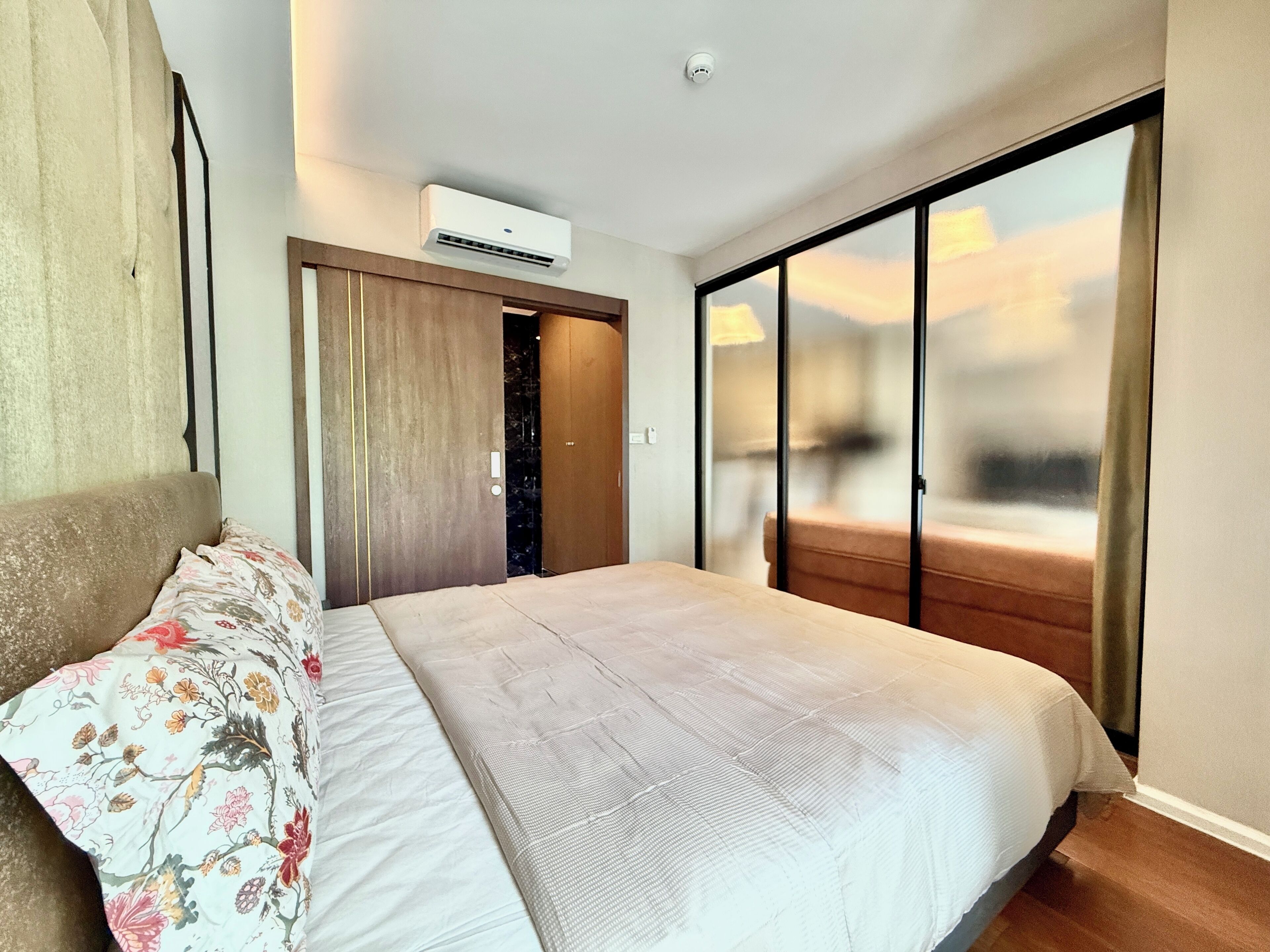 A hotel room with a bed, a wardrobe, an air conditioning unit, and a view through a large window.