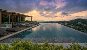 Une piscine dotée de chaises longues et d’un espace salon couvert, entourée de verdure et avec des bâtiments en arrière-plan.
