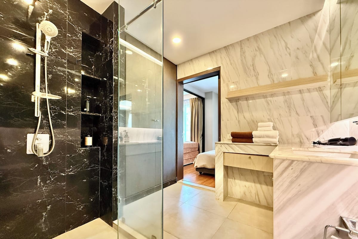 Ein modernes Badezimmer mit einer gläsernen Dusche, einem Marmorwaschtisch und einem sichtbaren Schlafzimmer mit Bett und Vorhängen.