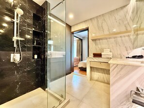 Een moderne badkamer met een douchecabine van glas, een marmeren wastafelmeubel en een zichtbare slaapkamer met bed en gordijnen.