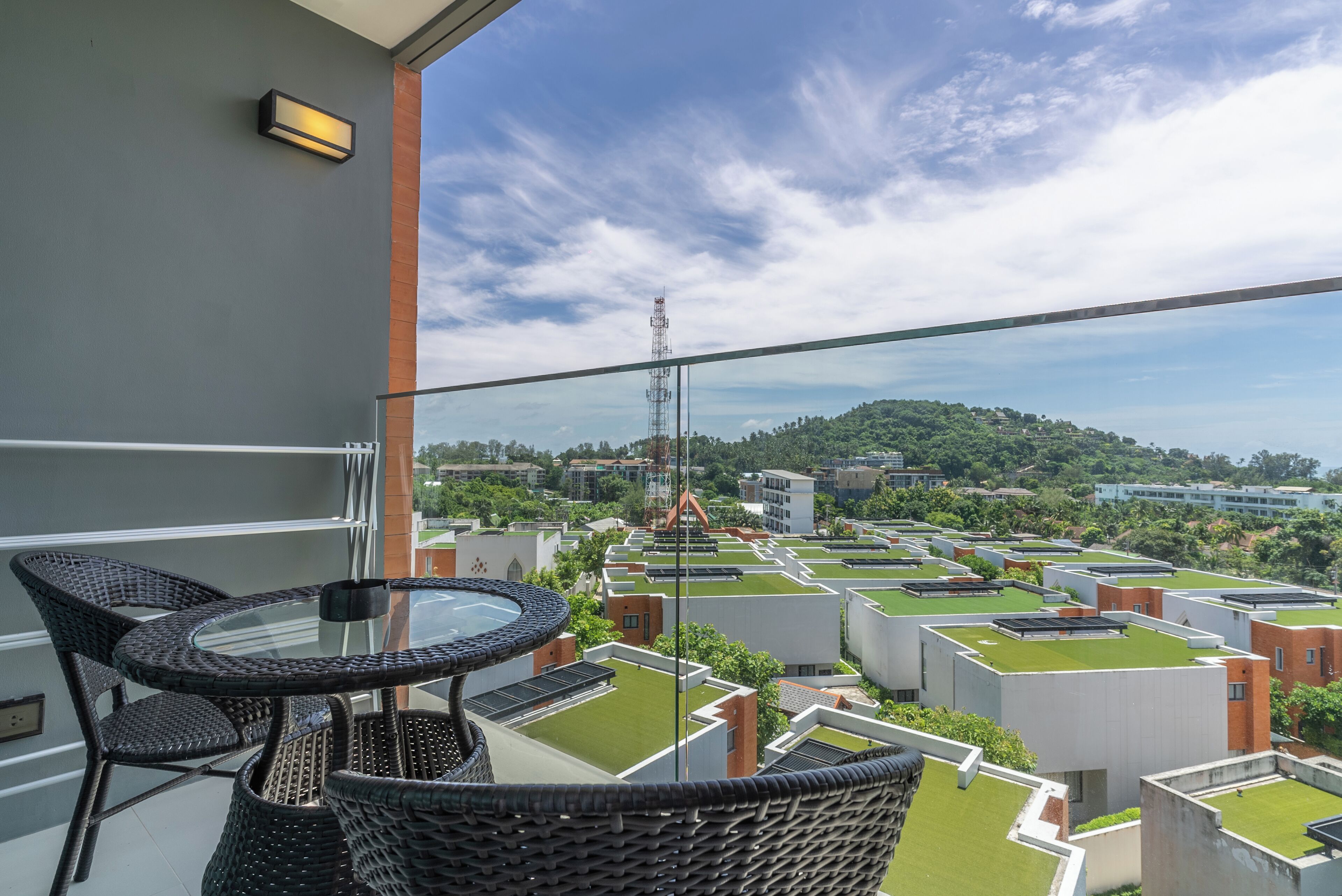 A balcony with a glass table and wicker chairs, overlooking a residential area with modern buildings and a green roof.