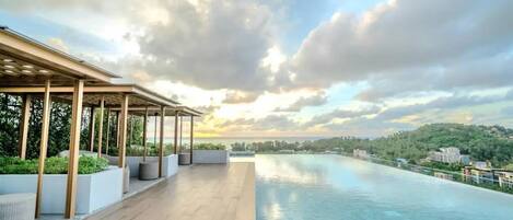A rooftop infinity pool with a wooden deck and outdoor seating area, overlooking a cityscape and hills.