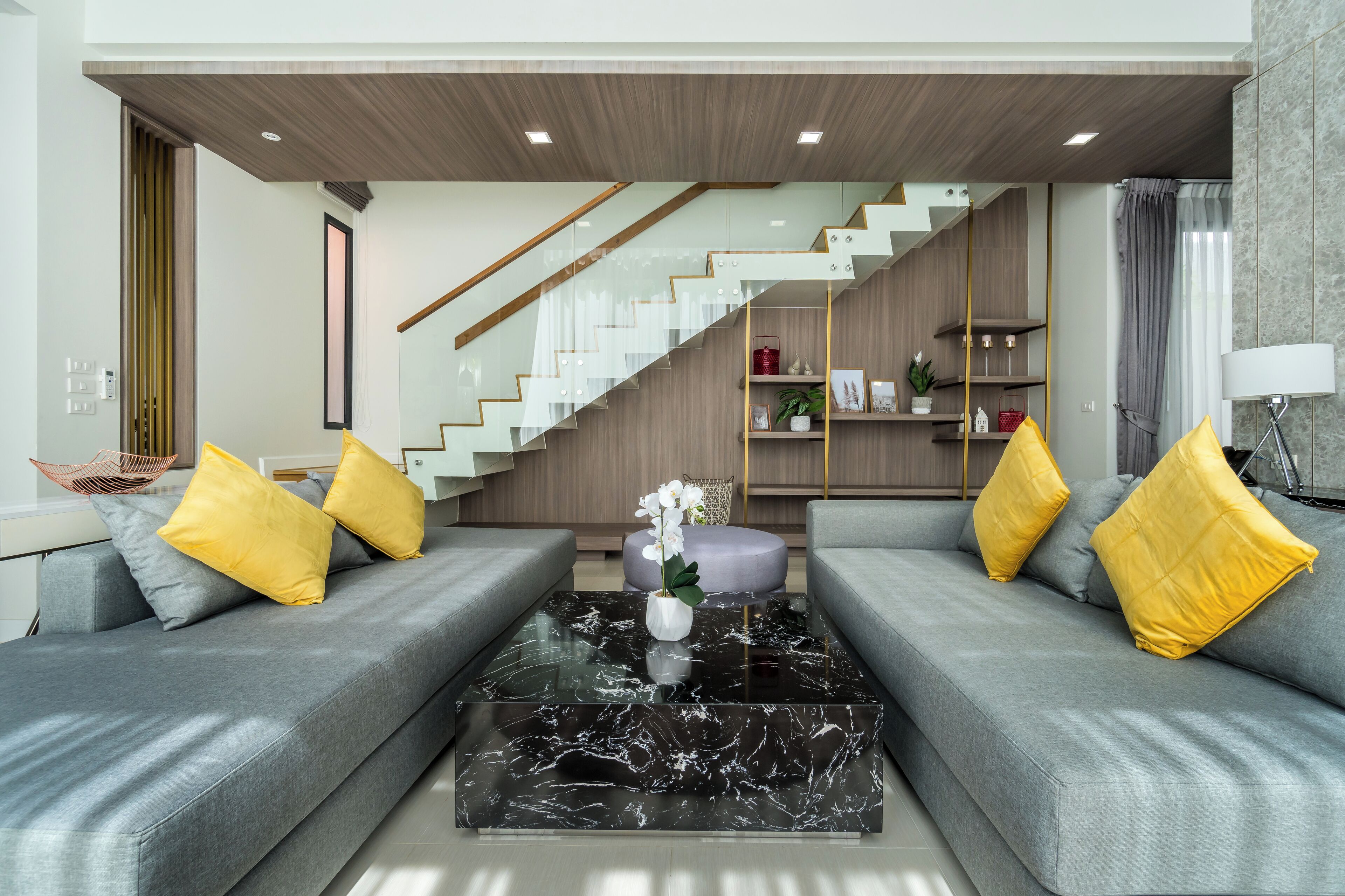 Un salon moderne avec des canapés gris, des coussins jaunes, une table basse en marbre et un escalier menant à une mezzanine en bois.