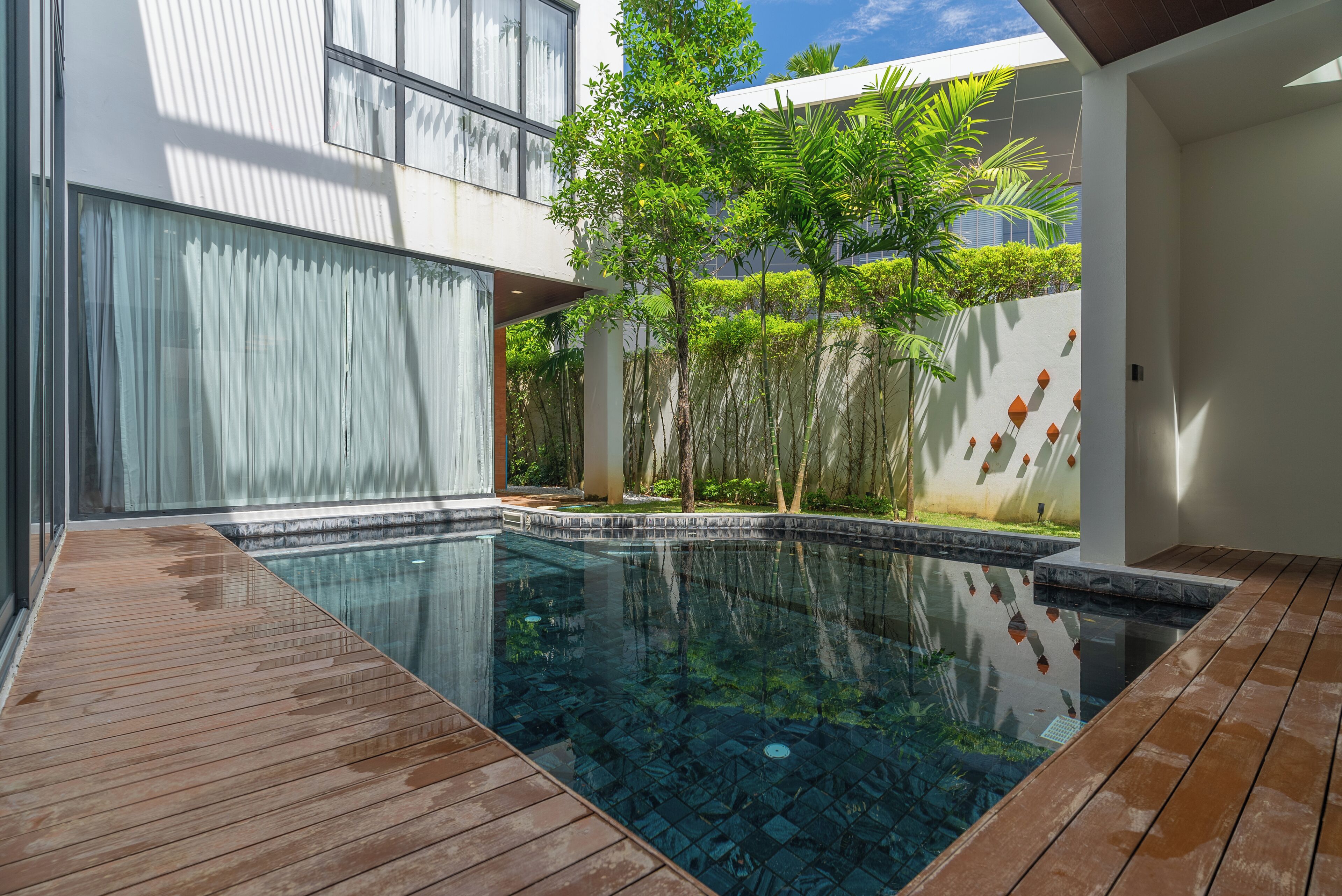 A modern outdoor pool area with a wooden deck, surrounded by lush greenery and a white building with large windows.