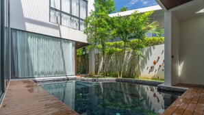 A modern outdoor pool area with a wooden deck, surrounded by lush greenery and a white building with large windows. - Panora Villa 8119 By Villacarte (Choeng Thale)
