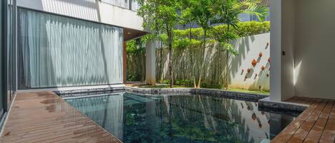 A modern outdoor pool area with a wooden deck, surrounded by lush greenery and a white building with large windows.