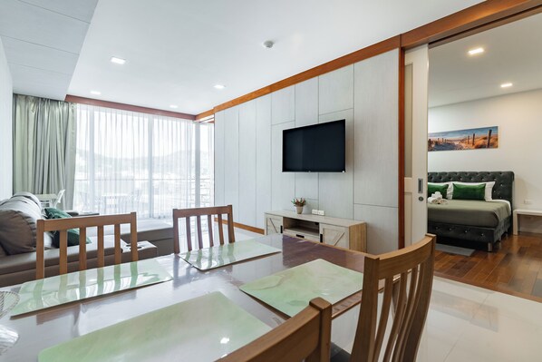 A modern living room with a dining area, a flat-screen TV mounted on the wall, and a cozy bed with a green pillow.