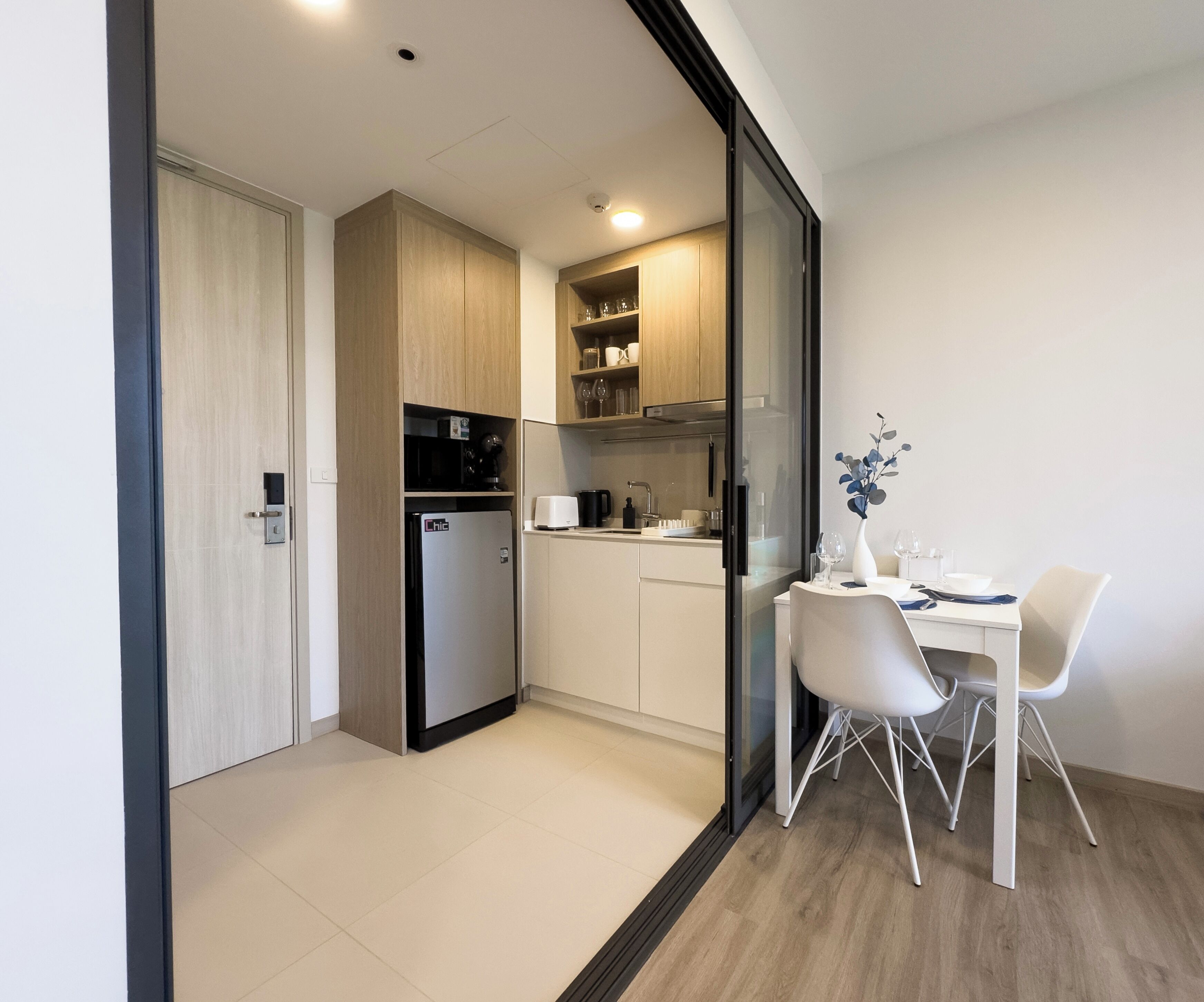 A modern kitchen with a built-in fridge, microwave, and coffee maker. There is a dining area with a white table and chairs, and a vase with flowers on the table.
