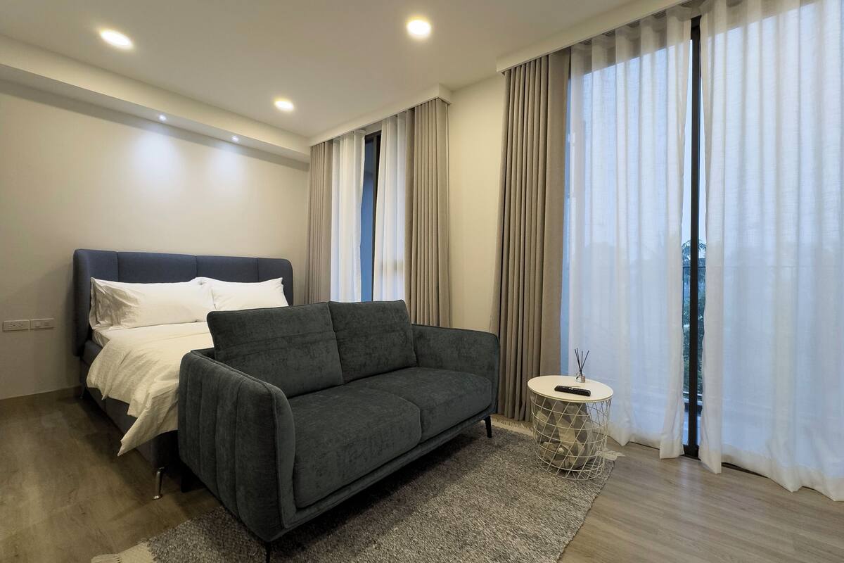 Une chambre d’hôtel moderne avec un canapé gris foncé, un lit avec une literie blanche, une petite table d’appoint ronde avec une télécommande, et de grandes fenêtres avec des rideaux légers.