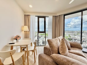 Una sala de estar moderna con un sofá, una mesa de comedor con sillas, una lámpara y vistas a un paisaje urbano a través de grandes ventanales.