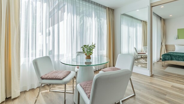 Une chambre d’hôtel moderne avec une table à manger en verre, deux chaises blanches, un lit recouvert d’une courtepointe verte et de grandes fenêtres ornées de rideaux légers.