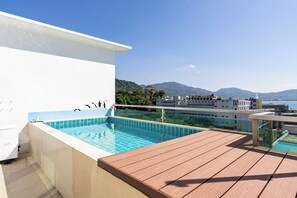 Et tagområde med pool, træterrasse, klart blåt vand og udsigt til bjerge og et vandområde i baggrunden.