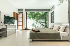 Un dormitorio moderno con cama, mesita de noche, televisor y vistas al verdor a través de un ventanal grande.