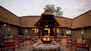 A circular stone fire pit surrounded by wooden chairs, with a thatched-roof building in the background. - ETNIKA MANOR HOUSE (Hoedspruit)