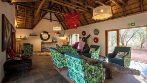 A spacious lounge area with a high ceiling and thatched roof, featuring green patterned sofas, a wooden bench, and a fireplace. - ETNIKA MANOR HOUSE (Hoedspruit)