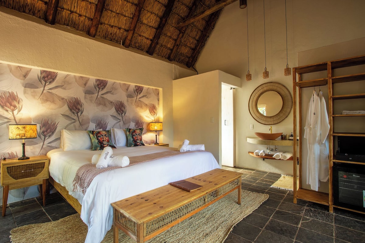 A bedroom with a bed, bedside tables, a wooden bench, a mirror, a wardrobe, and a bathroom with a sink and shelves.