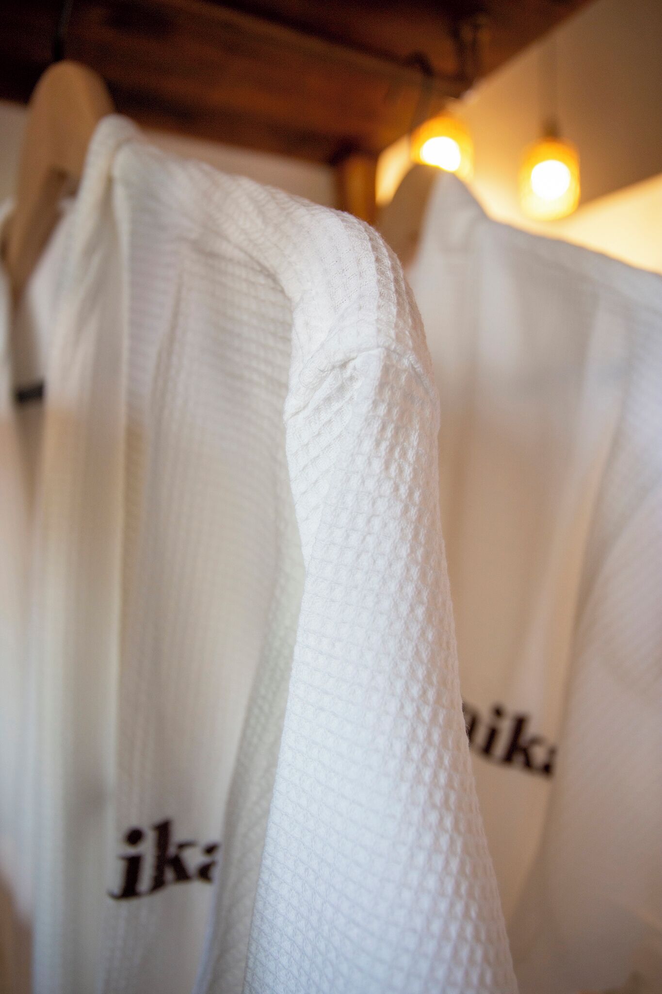 A close-up of white towels with the text "ika" printed on them, hanging in a room with wooden beams and warm lighting.