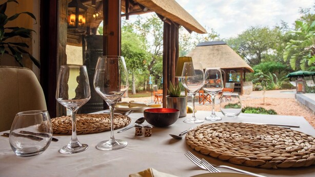 A dining table set with wine glasses, a woven placemat, and a bowl, with a view of an outdoor area and a thatched-roof structure. - ETNIKA MANOR HOUSE (Hoedspruit)