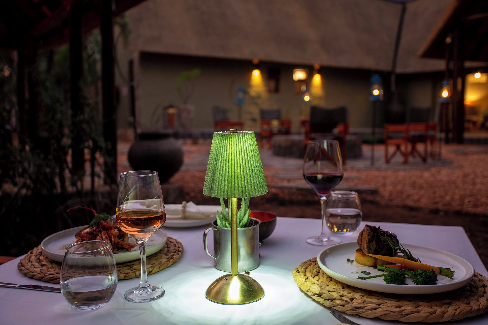 A dining table with plates of food, glasses of wine, and a green lamp.