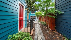 Un étroit sentier mène à une maison bleue avec une porte rouge, entourée de verdure et d’une clôture en bois.