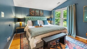 Une chambre à coucher avec une tête de lit capitonnée, une table de chevet avec une lampe, une grande fenêtre avec des rideaux et un tapis à motifs.
