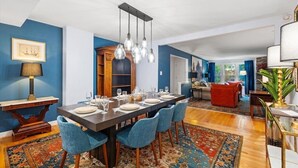 Un coin repas avec une longue table dressée pour six personnes, des chaises rembourrées de tissu bleu, un tapis à motifs et un luminaire suspendu.