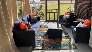 Une terrasse ensoleillée, meublée de meubles en osier, agrémentée d’un tapis coloré et offrant une vue sur une voiture et la verdure environnante.
