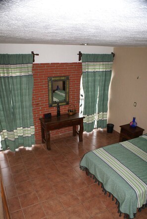 A bedroom with a brick wall, a wooden desk, a bed with a green bedspread, and green curtains. - Spacious 4-bedroom house in welcoming Oaxaca with beautiful terrace (Oaxaca de Juárez)