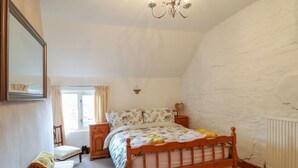 Ein Schlafzimmer mit einem hölzernen Bett, einem Nachttisch, einem Sessel, einem Fenster mit Vorhängen und einer weißen Ziegelwand.