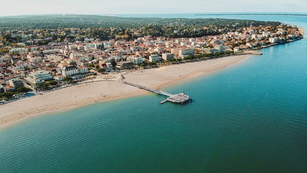 Vue aérienne d’une ville côtière avec une plage, une jetée et des bâtiments urbains denses.