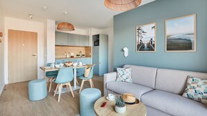 A modern living room with a grey sofa, blue chairs, and a wooden dining table. There are wicker lamps, a small table with a plant, and a wall-mounted TV.