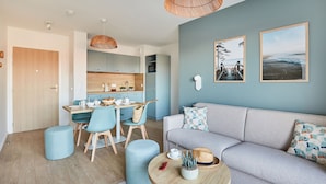 A modern living room with a grey sofa, blue chairs, and a wooden dining table. There are wicker lamps, a small table with a plant, and a wall-mounted TV.