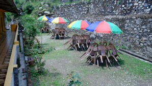 Outdoor seating with umbrellas, a stone wall, and a paved path. - Hotel Him Deep Uttarkashi (Bhatwari)