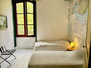 Een kamer met twee bedden, een raam met uitzicht op groen, een kaart aan de muur, een lamp en een stoel.