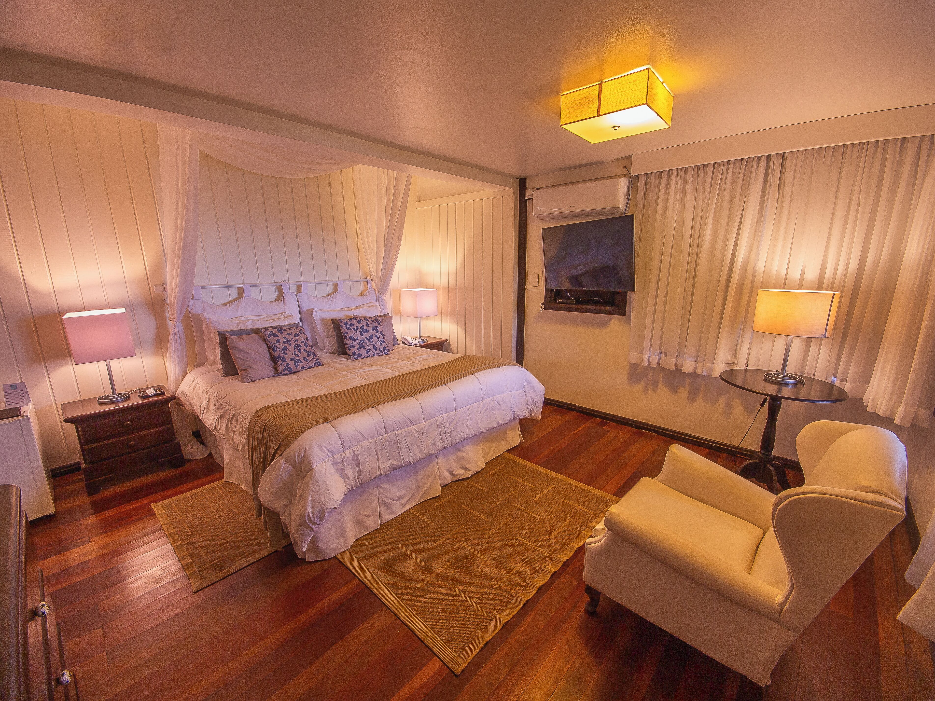 A hotel room with a bed, bedside tables, a chair, a small table, and a window with curtains.