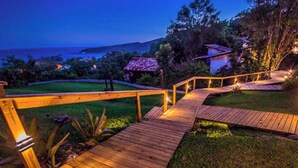 A wooden deck with a railing leading to a house surrounded by greenery and a view of the ocean. - Pousada Morada dos Bougainvilles (Imbituba)
