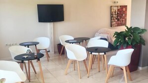Una sala moderna con un televisor de pantalla plana montado en la pared, un sofá y una mesa de centro. Hay varias sillas blancas y una planta en maceta.