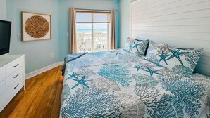 A bedroom with a bed, a ceiling fan, a window with a view, and a wall-mounted TV. - Shore Thing Premium Ocean View Two Bedroom Condo (Treasure Island)