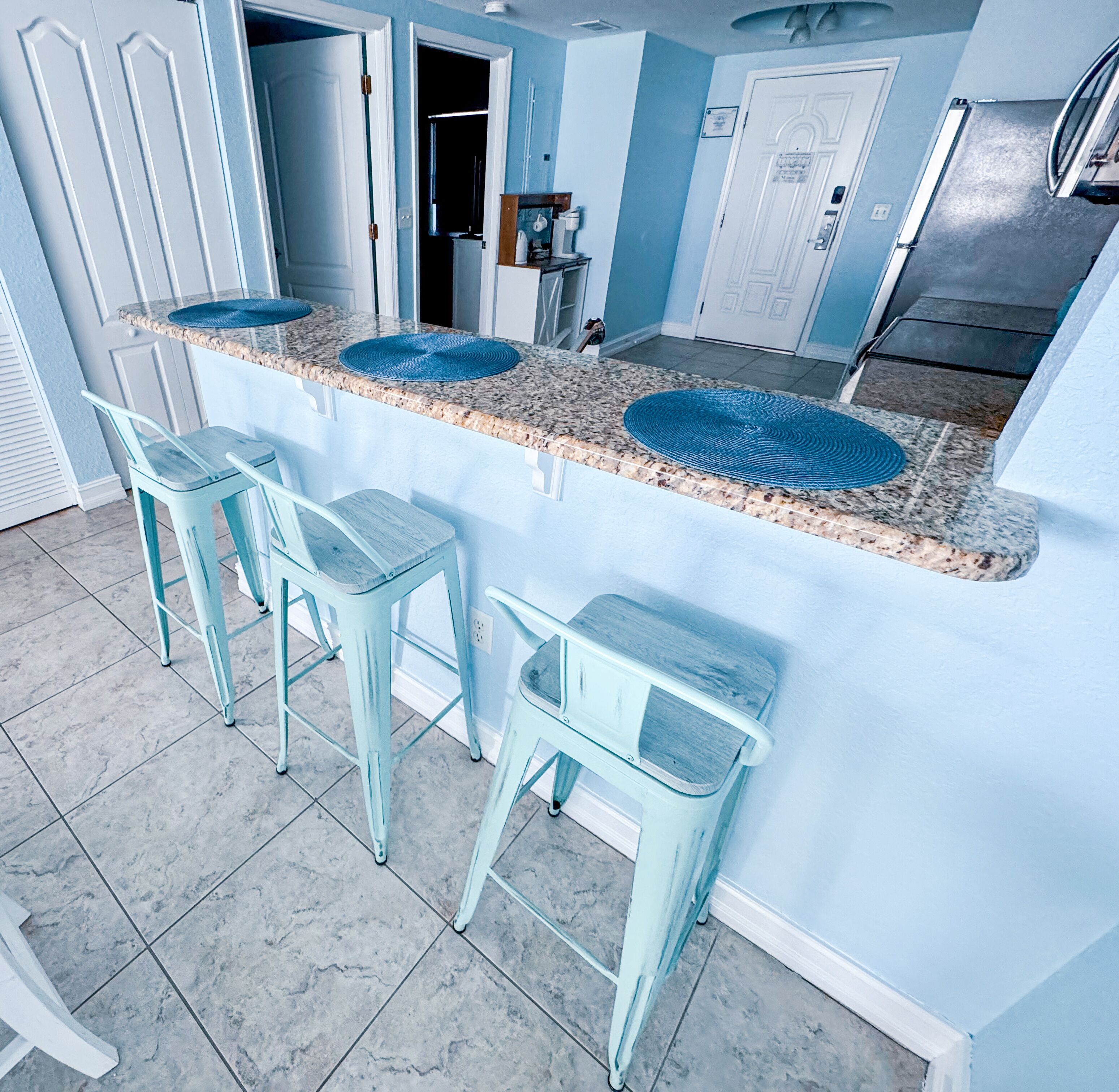 A kitchen with a granite countertop, two bar stools, and a stainless steel refrigerator.