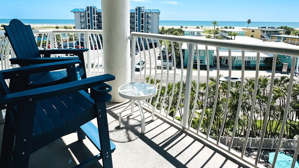 A balcony with a view of the ocean and buildings, featuring two Adirondack chairs and a small table. - Shore Thing Premium Ocean View Two Bedroom Condo (Treasure Island)
