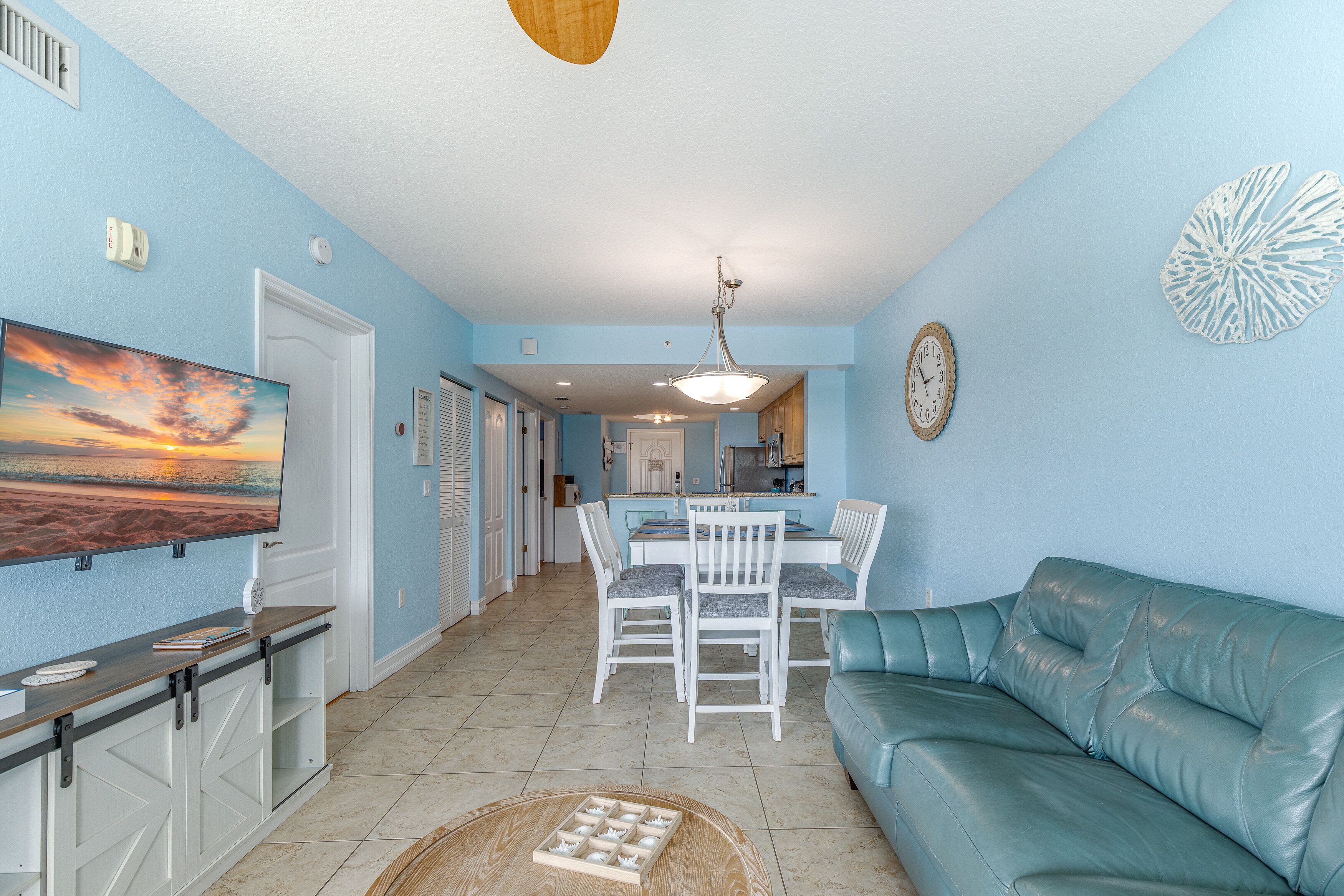 A living room with a blue couch, a dining table with chairs, a TV, and a kitchen in the background.
