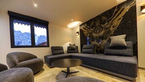 A modern living room with a sectional sofa, armchairs, a round coffee table, and a large window with a view of snow-covered trees. - Montaska - Chalet La Plagne - 7 Suites - Jacuzzi / Sauna (La Plagne Tarentaise)