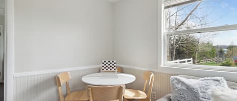 A corner room with a round table and four chairs, a window with a view, and a grey sofa with a fluffy pillow.