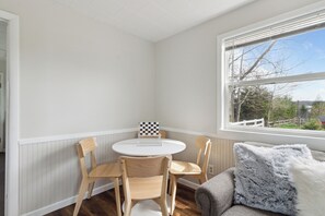 Ein Eckzimmer mit rundem Tisch und vier Stühlen, ein Fenster mit Aussicht und ein graues Sofa mit einem flauschigen Kissen.