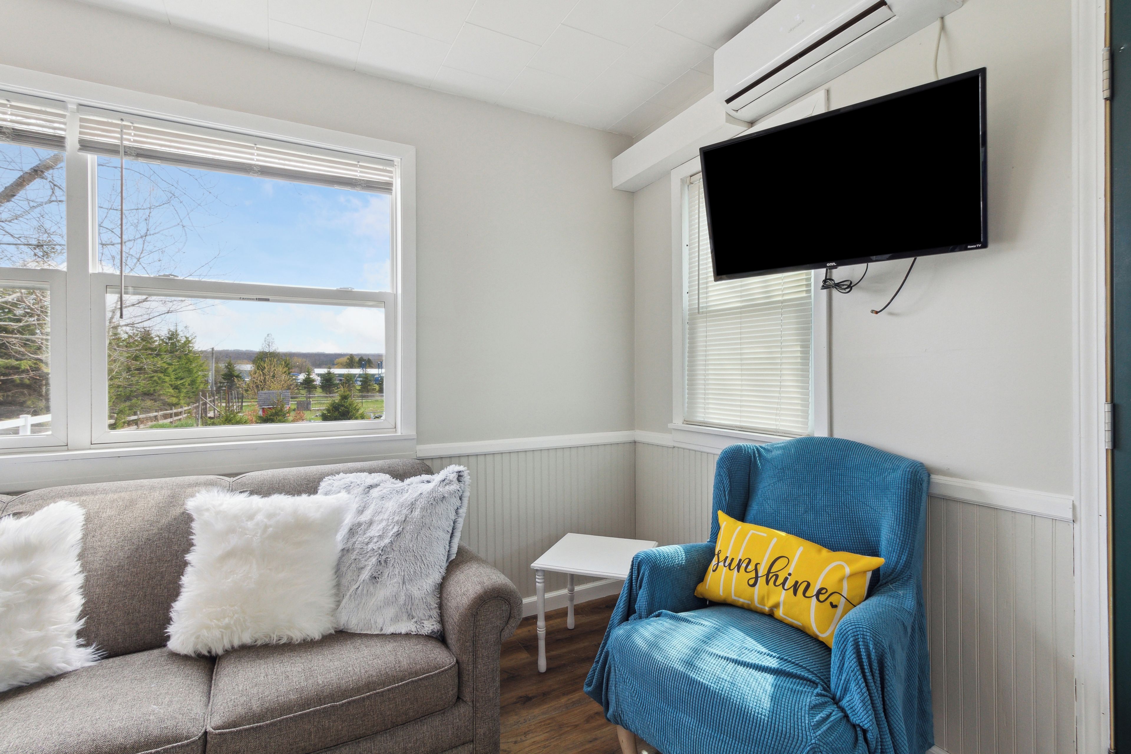 Una habitación con un sofá gris, un sillón azul con un cojín amarillo, una mesita blanca y un televisor montado en la pared.
