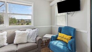 Una habitación con un sofá gris, un sillón azul con un cojín amarillo, una mesita blanca y un televisor montado en la pared.