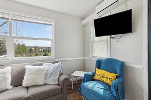 A room with a grey sofa, a blue armchair with a yellow pillow, a small white table, and a mounted TV.