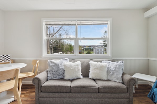 A living room with a gray sofa, two white fluffy pillows, a wooden dining table with chairs, and a window with a view of a house and trees.